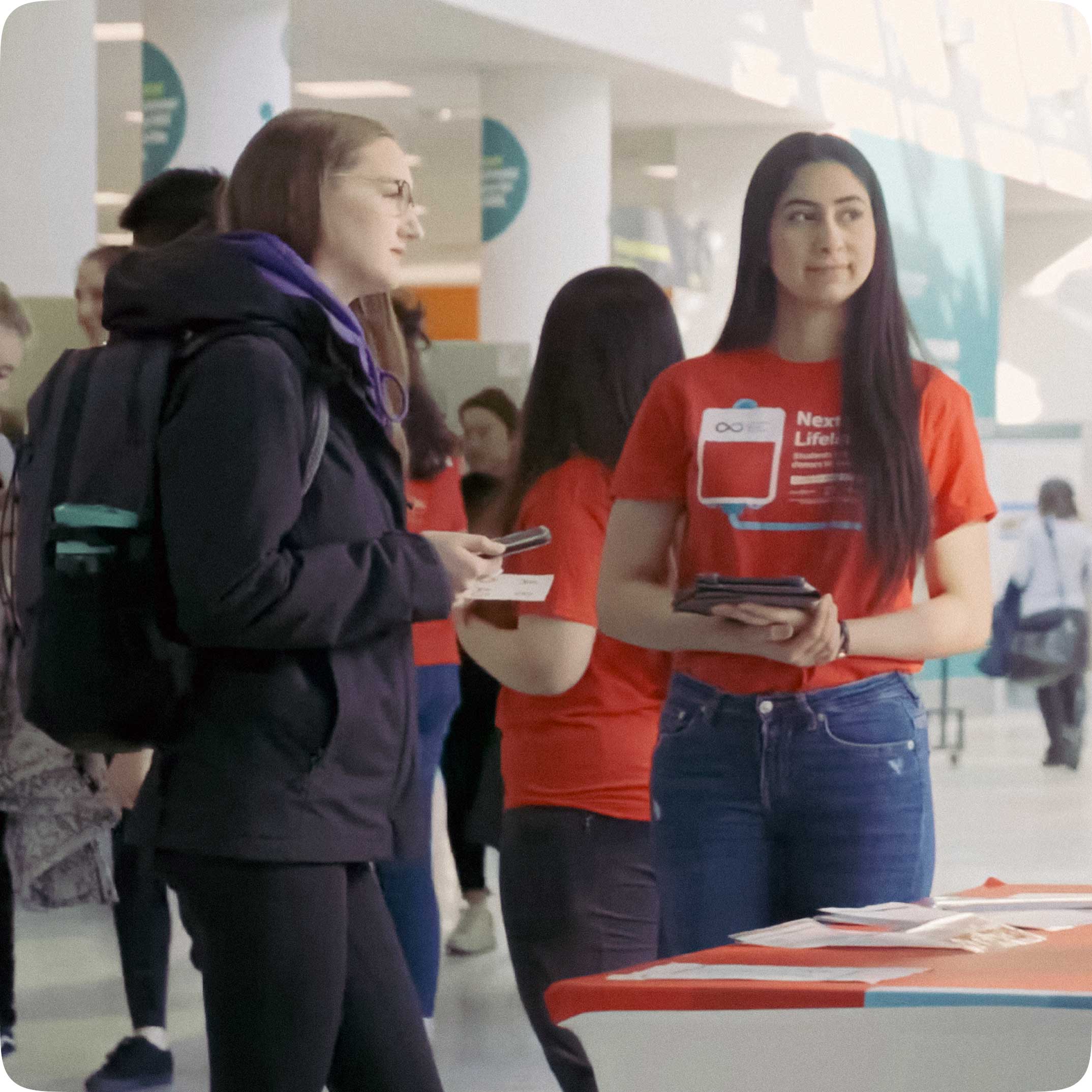 Une personne portant un sac à dos et un membre du personnel de la Société canadienne du sang sont debout devant une table sur laquelle est posé du matériel marketing. Elles regardent quelque chose sur le côté. En arrière-plan, il y a des gens et des tables.