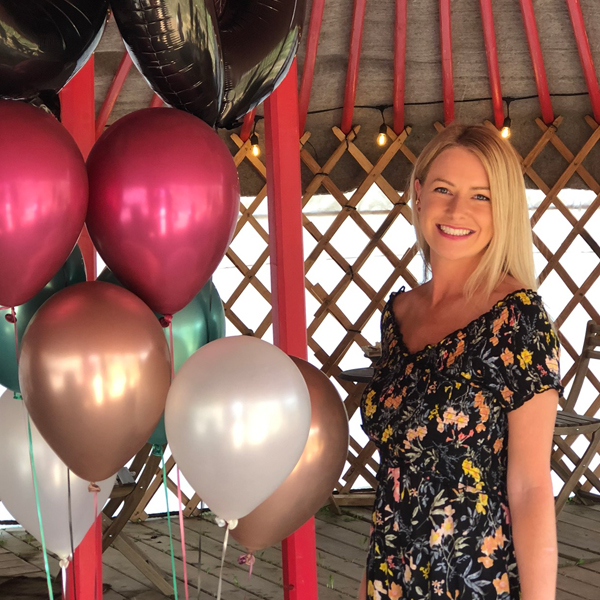 Image of blood donor Quinn Bleakley standing beside pink, teal, black, white, and gold balloons
