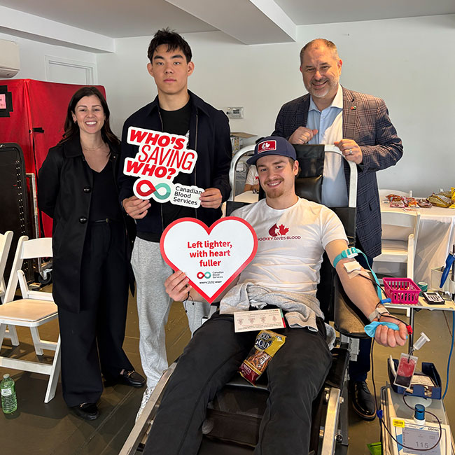Person donating in the chair holding up a heart shaped sign with 3 other people standing behind