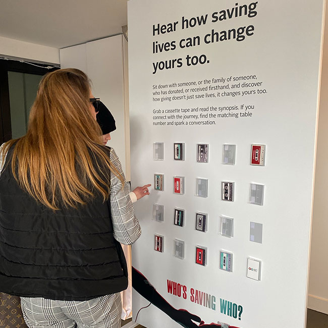 People looking at a large poster with the words Hear how saving lives can change yours too