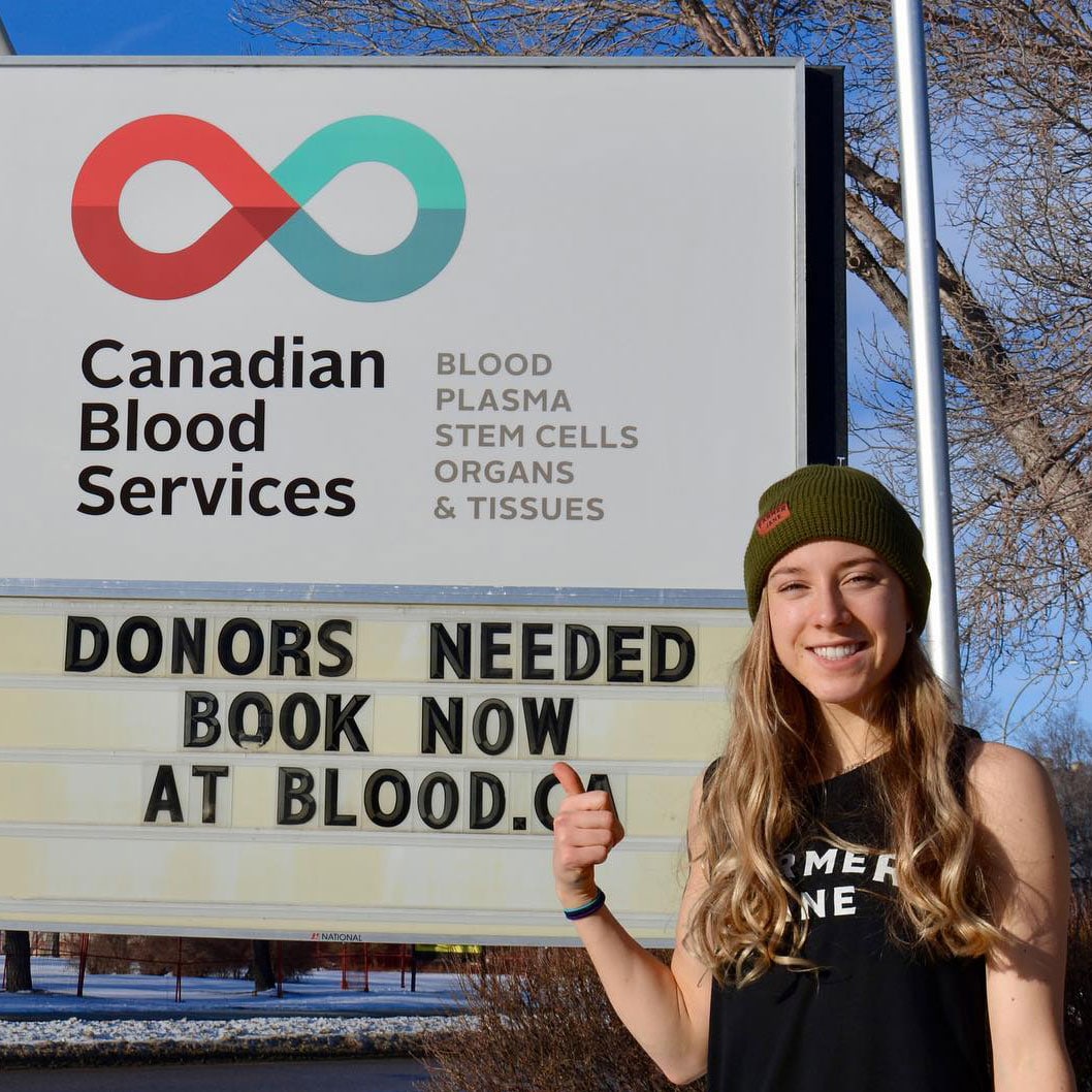 Une jeune femme se tient debout, près d’un panneau de la Société canadienne du sang, qui dit « Donors needed book now at blood.ca » (Nous avons besoin de donneurs maintenant à sang.ca)