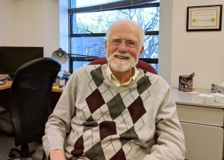 Dr. Michael McDonald sits in his office. He is wearing an Argyle sweater vest and has a white beard.