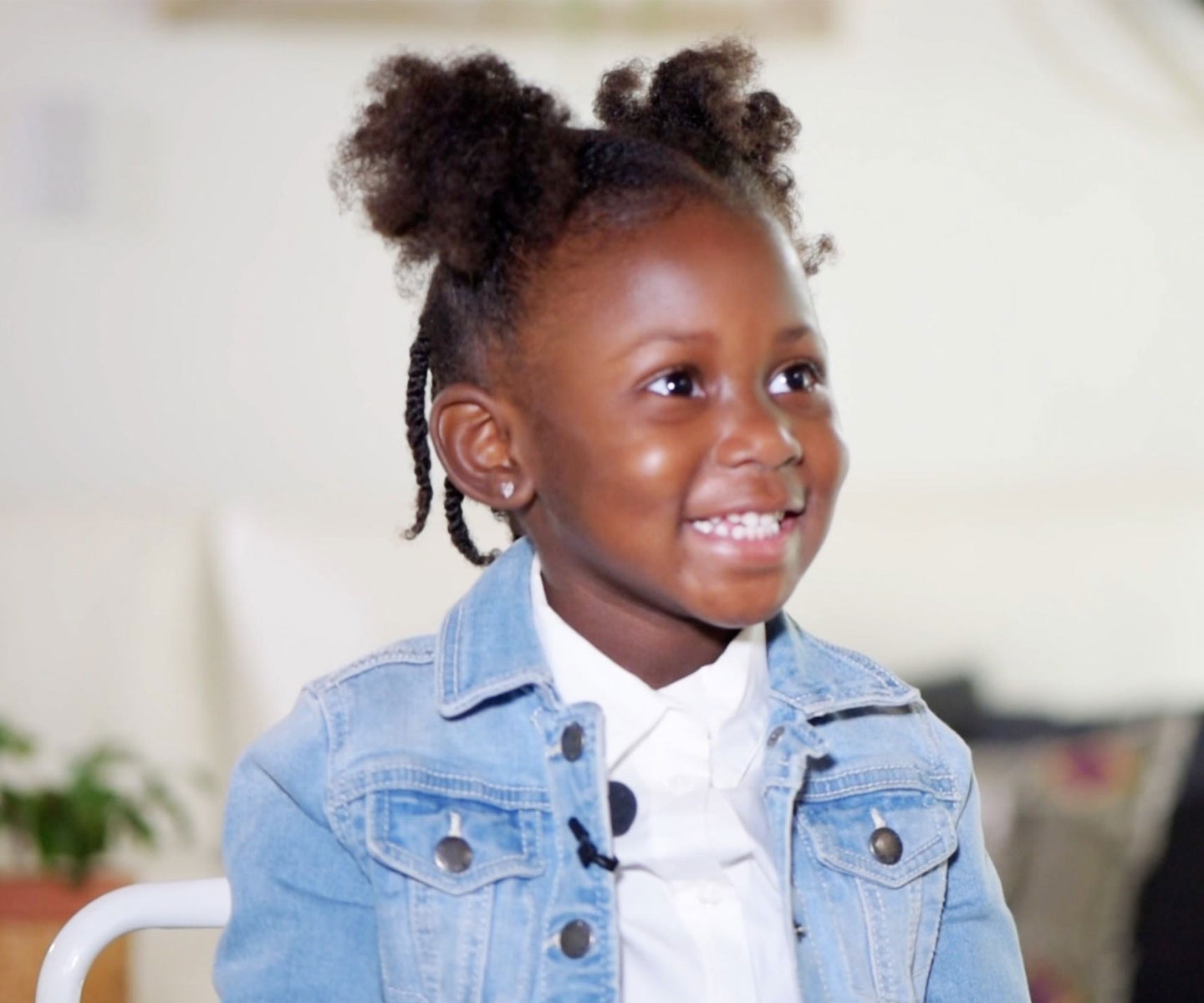 Petite fille d’âge préscolaire en veste en jean, assise en intérieur, regard hors champ et souriante