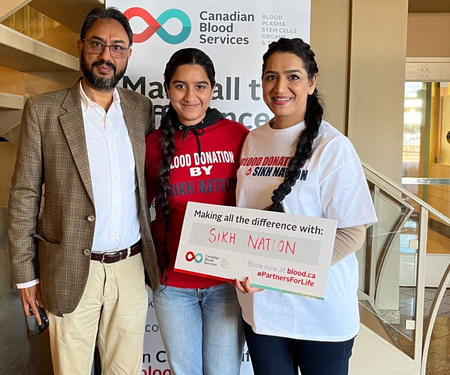 Une famille de trois personnes se tient ensemble derrière une pancarte sur laquelle est inscrit « Making all the difference with: Sikh Nation ». Deux d’entre elles portent un t-shirt Blood donation by Sikh Nation.