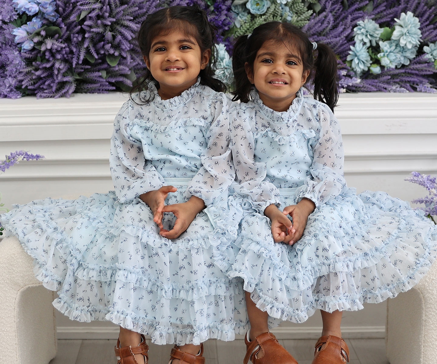 Identical twin girls in blue dresses seated near purple flowers