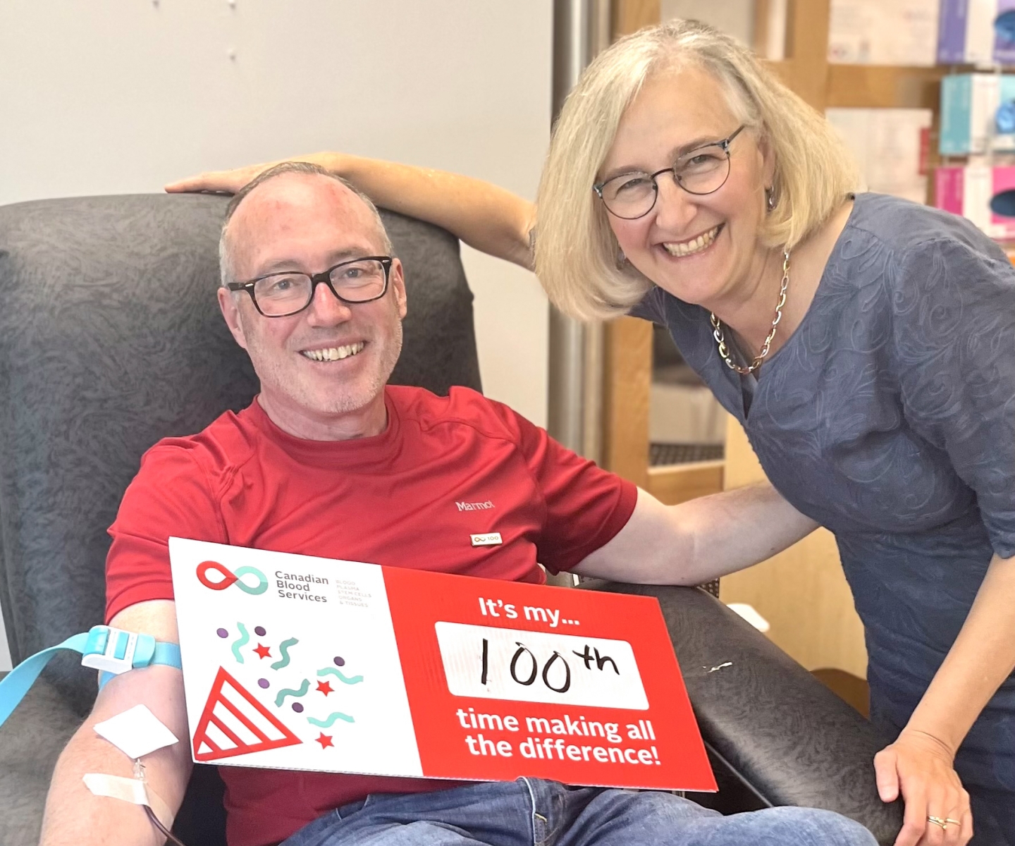 Blood donor in donor bed holding sign that says "It's my 100th time making all the difference" with wife beside him