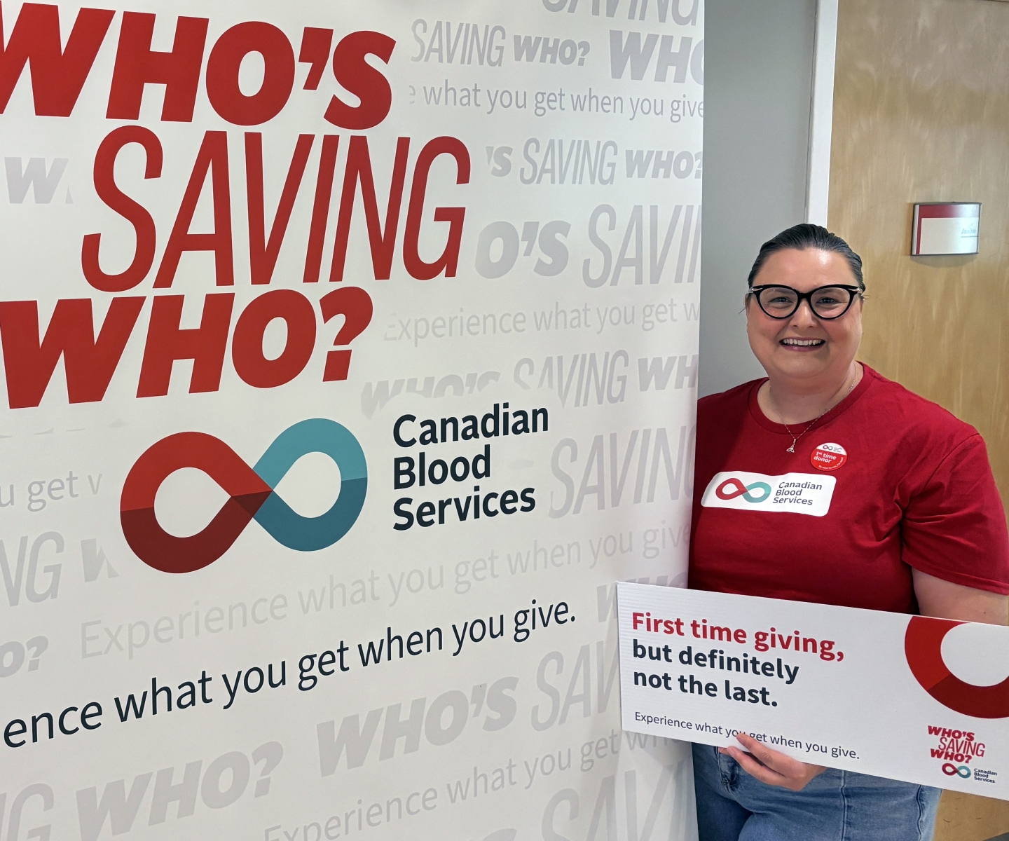 Blood donor in red shirt next to sign that say's Who's Saving Who?