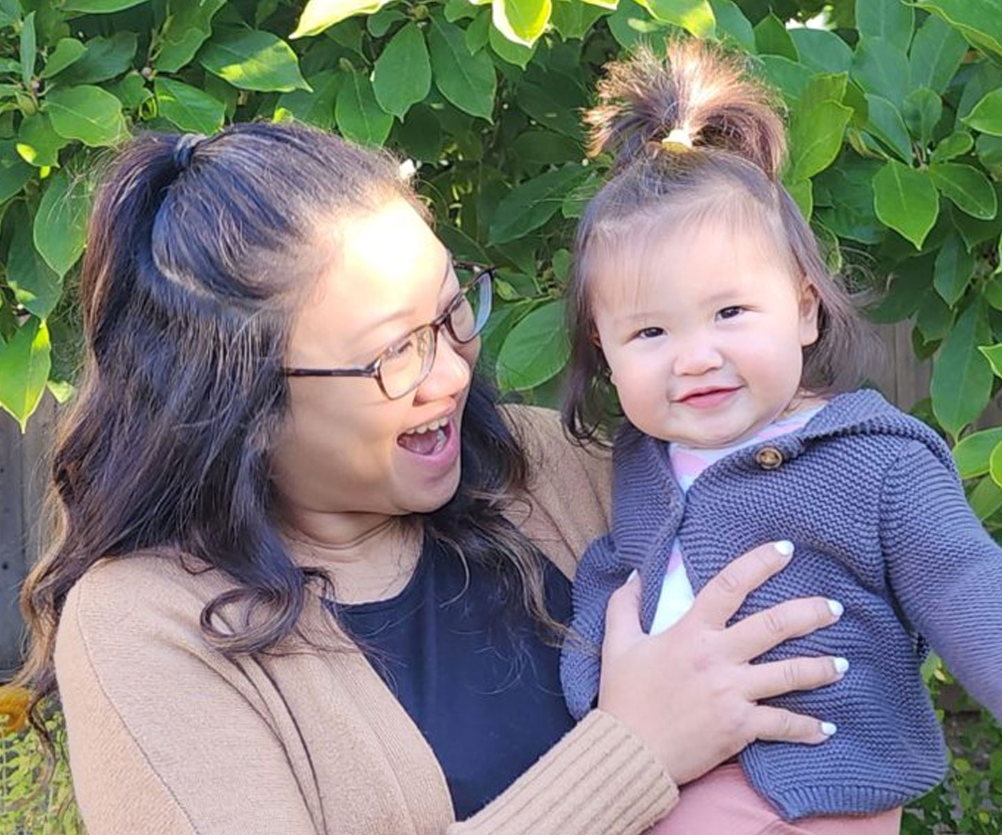 Smiling mother, holding a toddler, standing in front of a tree.