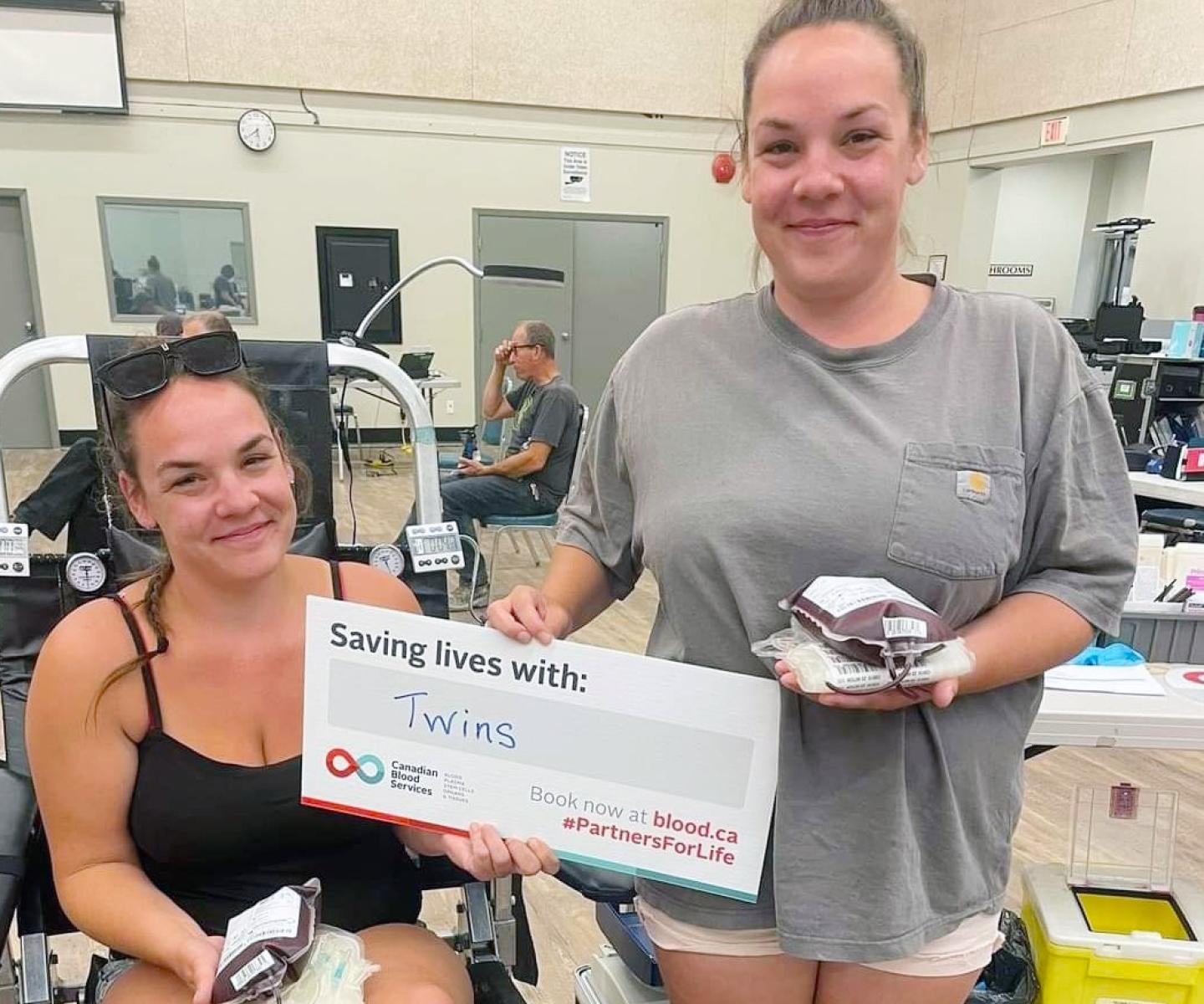 Twin blood donors holding blood donations and Canadian Blood Services sign that says Saving lives with: Twins.