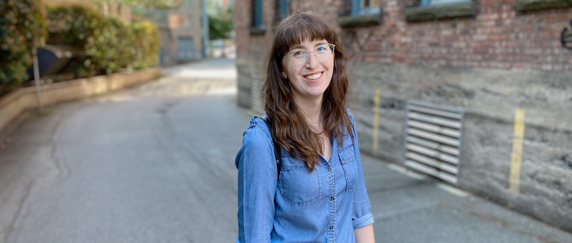 A woman with long brown hair, gold-rimmed glasses and a denim shirt stands on a roadway lined with greenery and brick buildings. 