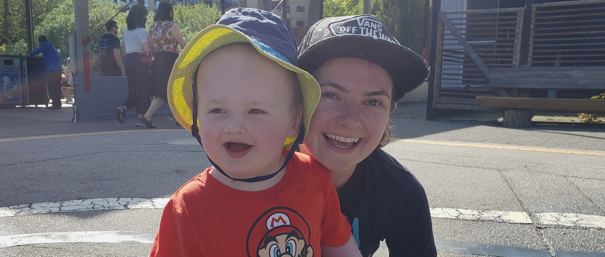 Mother crouched down on paved road with arm around the waist of her 2-year-old son and passersby in the background