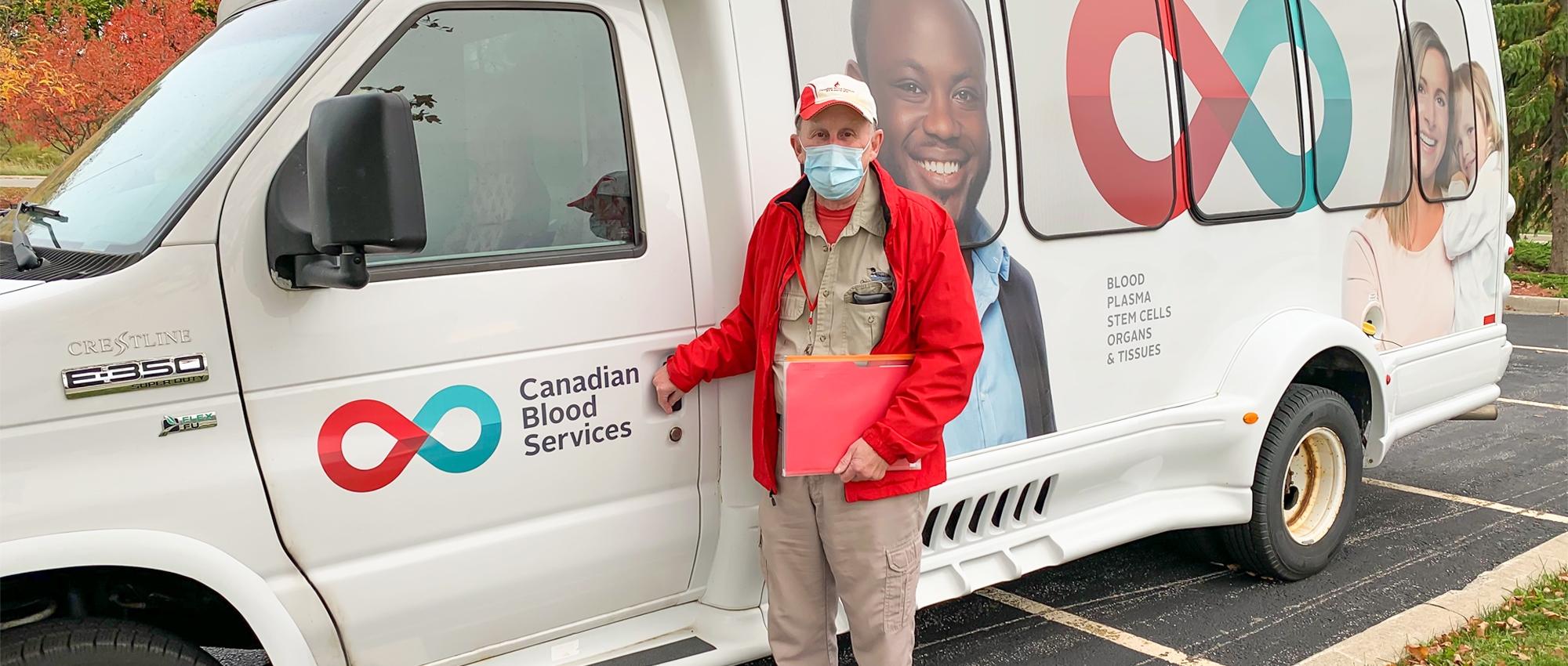 Image of Volunteer bus driver David Neu wearing a mask and holding a clipboard stands at the driver-side door of a short white bus with the Canadian Blood Services logo.