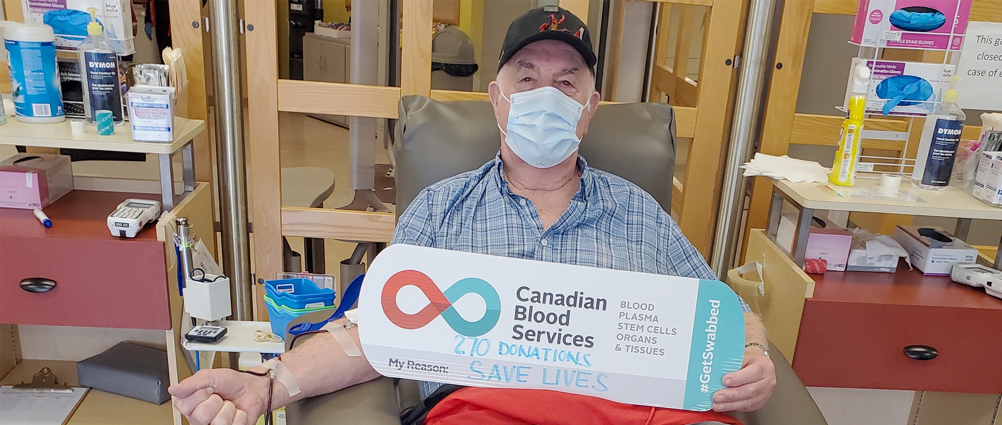 An 87-year-old man who is donating blood holds a sign that says “270 donations. My reason? Save lives.”