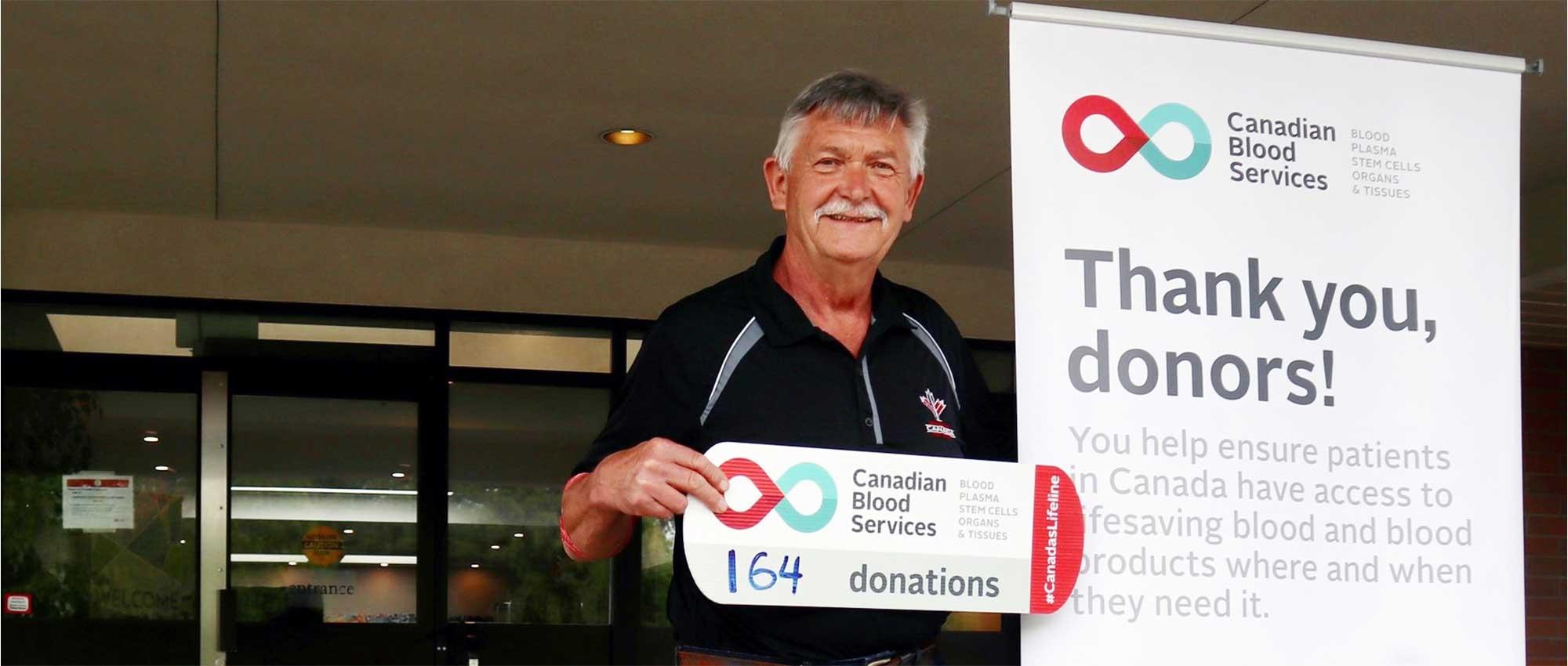Un donneur de sang se tient debout, à côté d’un panneau qui dit, en anglais, « merci aux donneurs ».