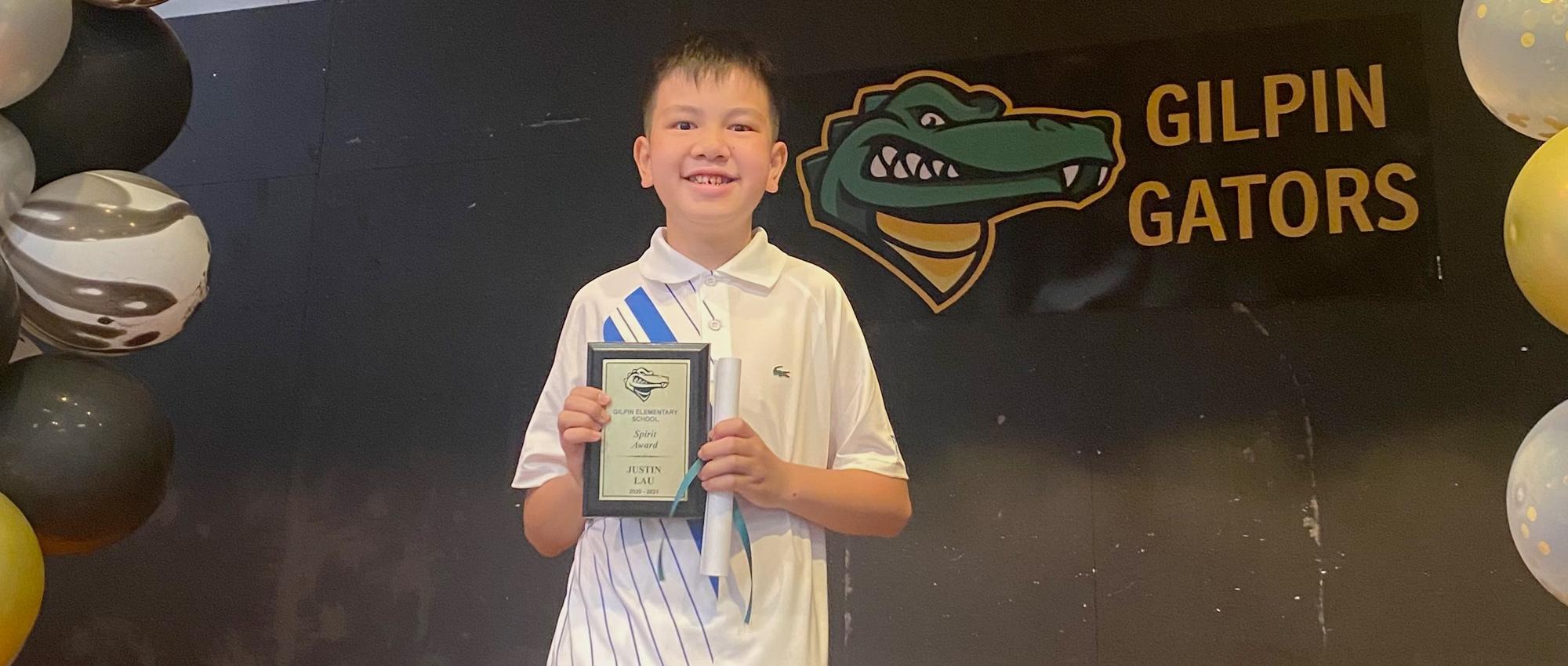 An Asian boy smiles as he holds his award plaque in front of a black background with “Gilpin Gators” inscription and logo. 