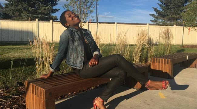 Revee Agyepong seated outdoors on a bench, leaning back on her right arm, legs crossed and smiling with a wood fence in the background