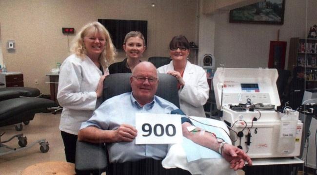 Man sits in blood donor chair holding a sign that says 900, surrounded by staff