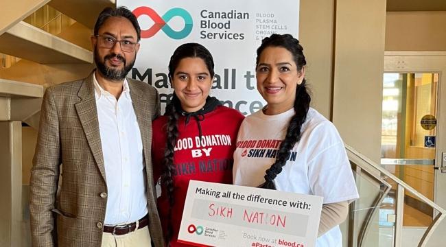 Une famille de trois personnes se tient ensemble derrière une pancarte sur laquelle est inscrit « Making all the difference with: Sikh Nation ». Deux d’entre elles portent un t-shirt Blood donation by Sikh Nation.