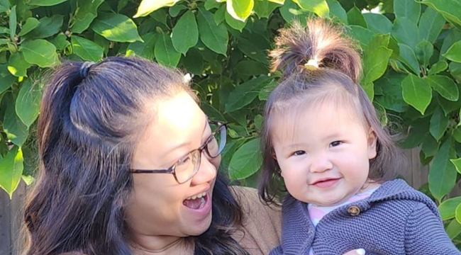 Smiling mother, holding a toddler, standing in front of a tree.