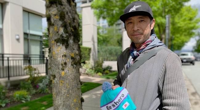 man, in a baseball cap, stands by a tree holding a toque with ‘be a hero’ written across the front