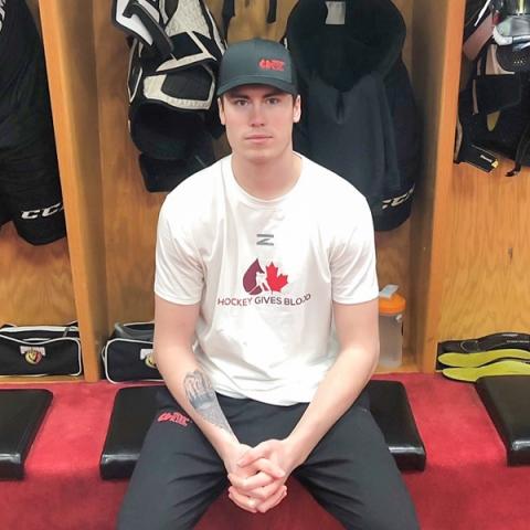 Blood donor Carter Robinson sitting in a hockey locker room wearing a Hockey Gives Blood white t-shirt with a black cap