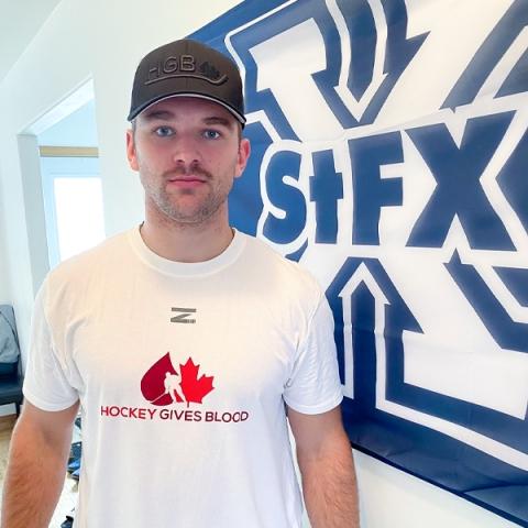 Image of blood donor Ethan Crossman in a white Hockey Gives Blood t-shirt by a wall with the letters StFX