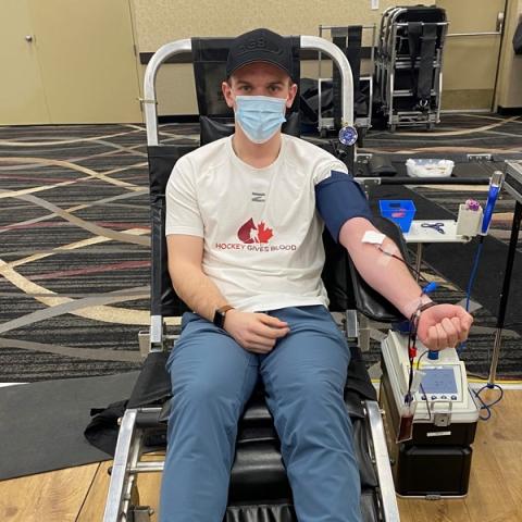Image of Trent Miner sitting in the donation chair donating blood at the donor centre wearing a black baseball cap and white hockey gives blood t-shirt