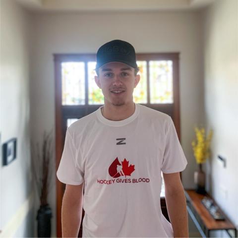 Image of blood donor Jake Chiasson wearing a black cap and a white hockey gives blood t-shirt standing in an inside entryway