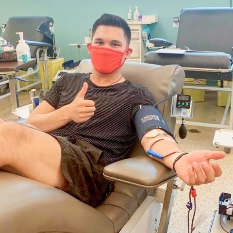 Image of Jett Woo sitting in a chair donating blood giving a thumbs up at the donor centre wearing a dark grey t-shirt and shorts.
