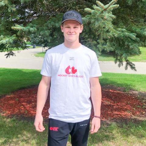 Image of blood donor Reid Valade wearing a white Hockey Gives Blood t-shirt standing out in front of a pine tree.
