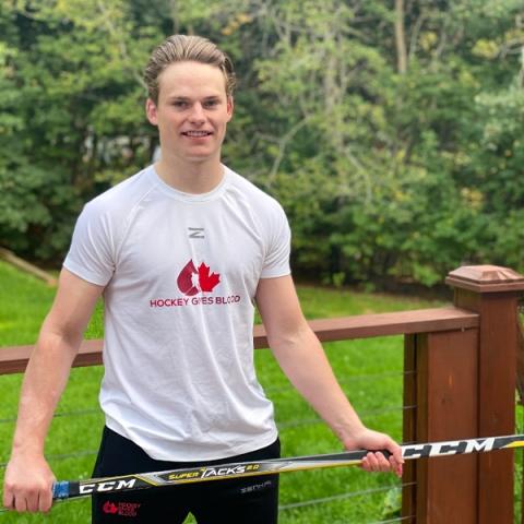 Blood donor Thomas Casey wearing a hockey gives blood t-shirt holding a hockey stick in both hands standing outside backyard